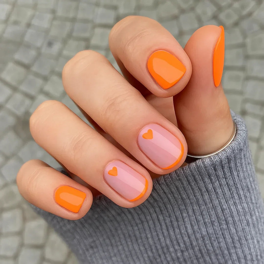 Hand with short nails featuring orange French tips and heart nail art design
