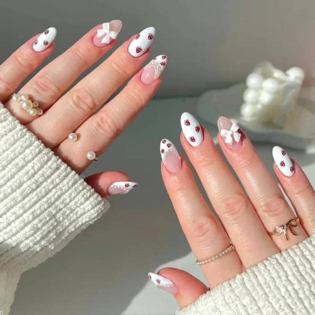 White 3D bow and strawberry nail art on almond-shaped nails with pearl rings
